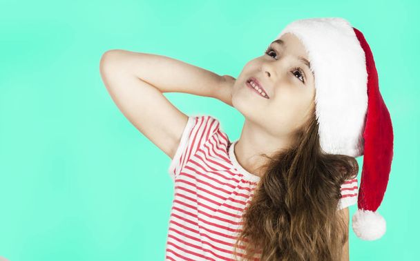 Little bright beautiful girl emotionally posing in Santa hat on an isolated turquoise background - Photo, image