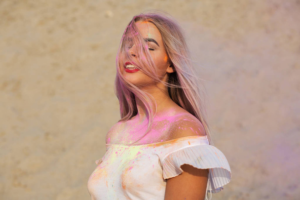 Closeup shot of glorious blonde woman with red lipstick covered with pink dry paint Holi at the desert - Фото, изображение