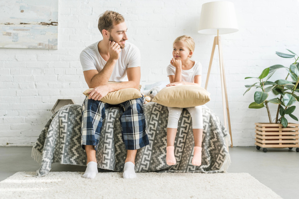 happy father and cute little daughter sitting with pillows on bed and smiling each other - Foto, afbeelding