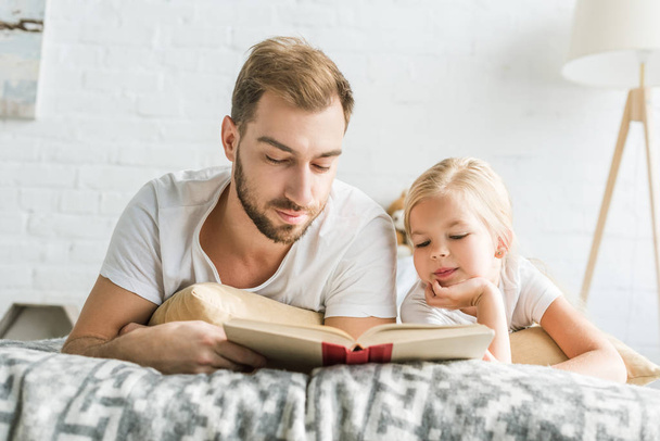 father and adorable little daughter lying on bed and reading book  - Фото, зображення