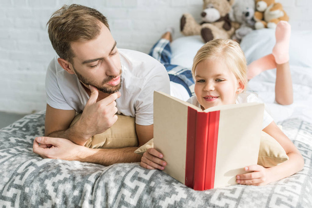 father looking at cute little daughter reading book on bed - Fotoğraf, Görsel