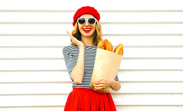 Portrait happy surprised laughing woman wearing red beret holding paper bag with long white bread baguette on white wall background - Fotografie, Obrázek