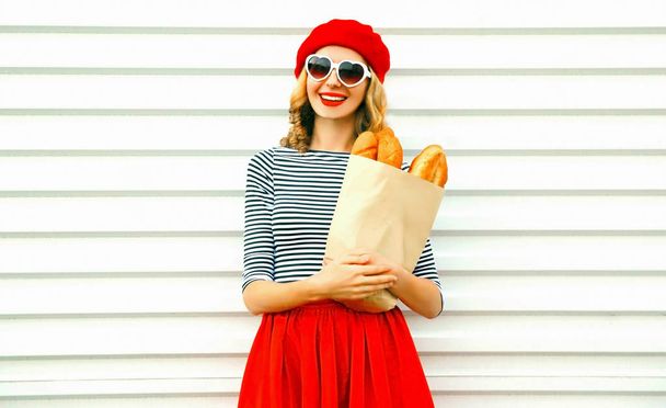 Portrait happy cute smiling woman wearing red beret holding paper bag with long white bread baguette on white wall background - Fotografie, Obrázek