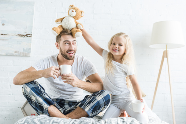 милая маленькая дочь играет с плюшевым мишкой и улыбается в камеру, пока счастливый отец пьет чай на кровати
 - Фото, изображение
