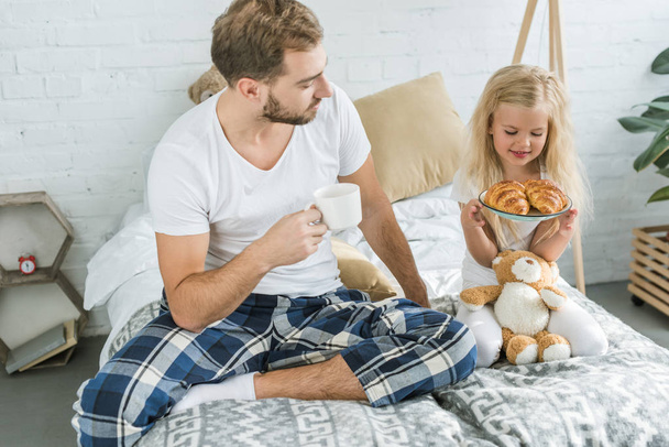 Vater hält Tasse Kaffee und schaut kleine Tochter mit leckeren Croissants auf dem Teller an  - Foto, Bild