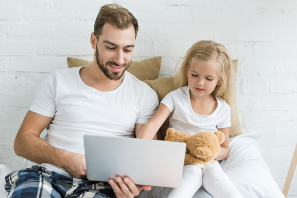 happy father and cute little daughter with teddy bear using laptop together in bedroom   - Foto, Imagen