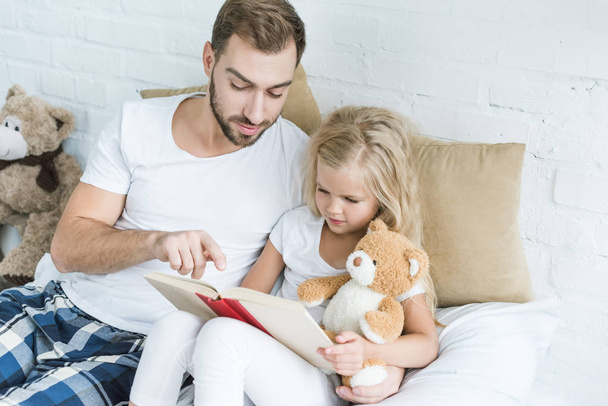 high angle view of father and adorable little daughter reading book together on bed - Fotoğraf, Görsel