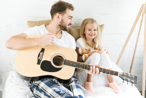 щасливий батько з акустичною гітарою дивиться на милу маленьку доньку з плюшевим ведмедем на ліжку
 - Фото, зображення