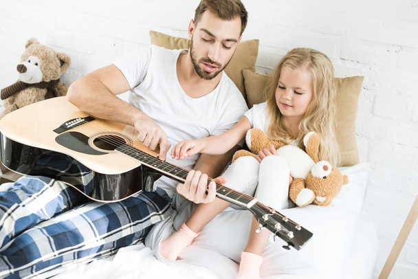 high angle view of father and cute little daughter with teddy bear looking at acoustic guitar on bed  - Photo, Image
