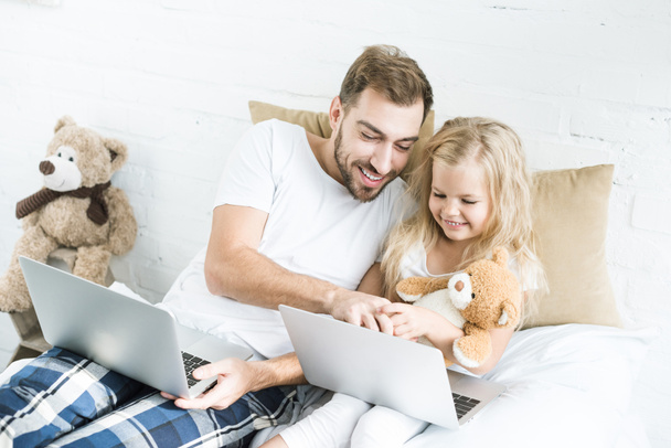 happy father and daughter using laptops on bed - Фото, изображение
