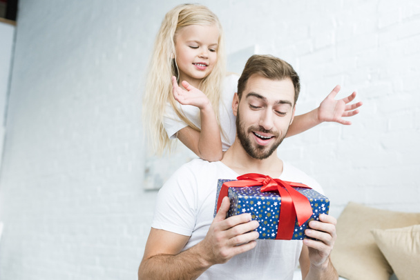 happy little daughter looking at surprised father holding gift box at home  - Фото, зображення