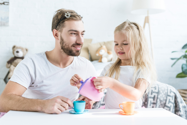 happy father looking at smiling little daughter pouring tea from toy kettle - Photo, Image
