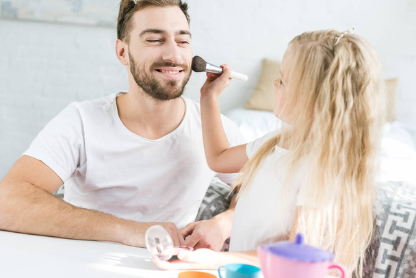 adorable little daughter applying makeup to happy bearded father at home - Photo, Image