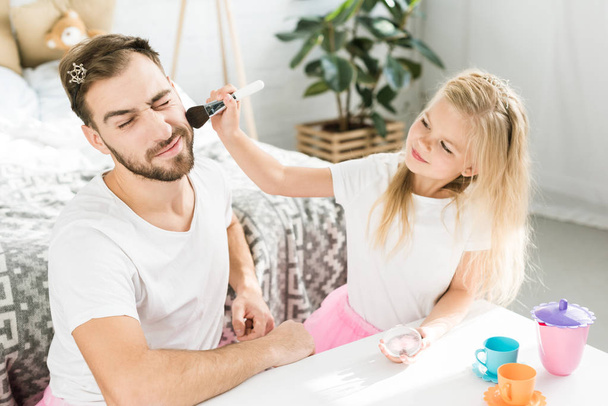 smiling little daughter applying makeup to bearded father at home - Foto, Imagen