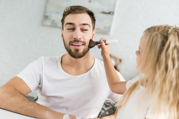 little daughter applying makeup to smiling bearded father at home - Foto, immagini