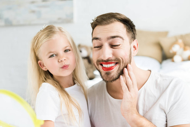 adorable little daughter and happy bearded father with red lipstick having fun together at home - Foto, afbeelding