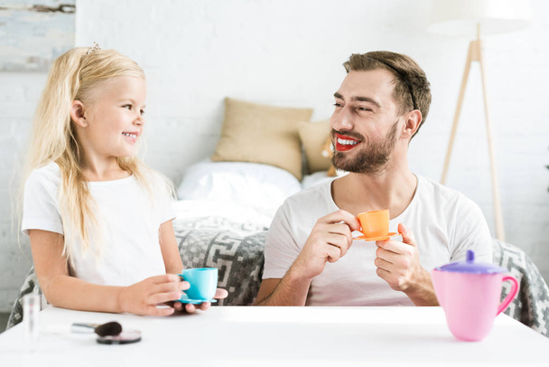 father with red lipstick and cute happy daughter smiling each other while playing with toy dishes at home  - Photo, Image