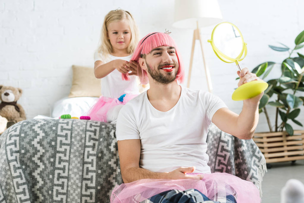 adorable córeczka w spódnicy tutu bawić szczęśliwy ojciec w różowych peruk, patrząc w lustro      - Zdjęcie, obraz