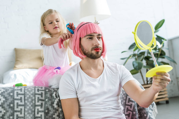 adorable little daughter in tutu skirt playing with father in pink wig looking at mirror     - Foto, Bild