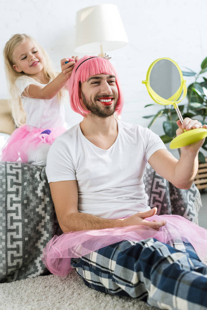милая маленькая дочь в юбке в пачке, играющая с улыбающимся отцом в розовом парике, смотрящим в зеркало
    - Фото, изображение