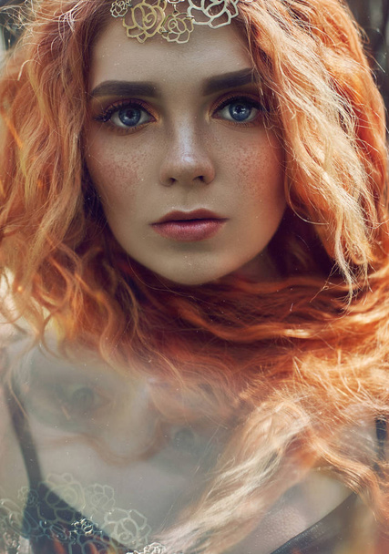 Beautiful redhead Norwegian girl with big eyes and freckles on face in the forest. Portrait of redhead woman closeup in nature, fabulous mysterious appearance long wavy red hair in the sun. Hair care - 写真・画像