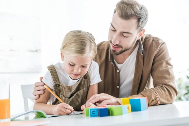 father looking at cute little daughter writing in workbook and learning at home - Foto, Bild