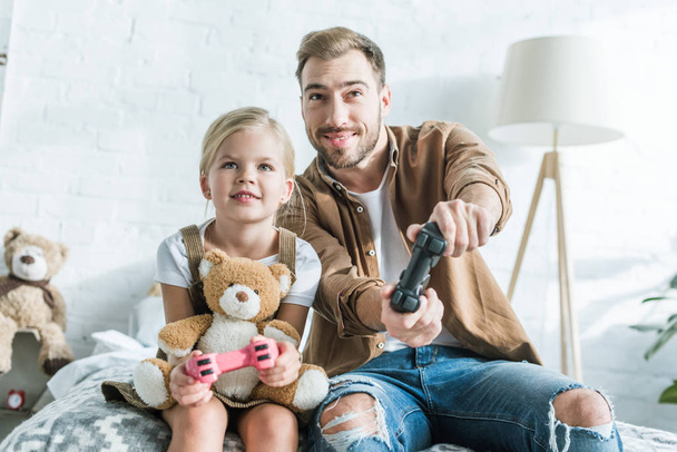 feliz padre y linda hijita con oso de peluche jugando con joysticks en casa
 - Foto, imagen