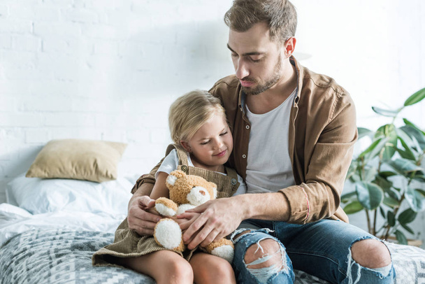 father looking at cute little daughter sitting with teddy bear on bed  - Foto, imagen