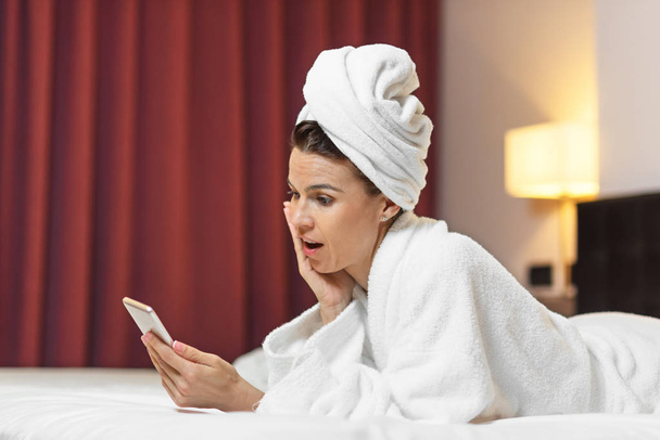 Νεαρή γυναίκα στο μπουρνούζι ξαπλωμένος σε δωμάτιο ξενοδοχείου, χρήση κινητού τηλεφώνου, χαλαρή μετά το μπάνιο - Φωτογραφία, εικόνα
