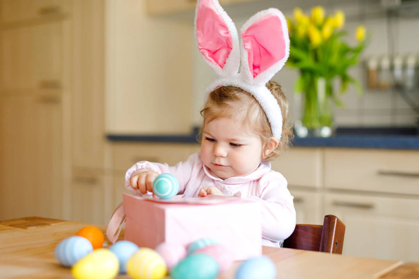 Linda niña pequeña con orejas de conejo de Pascua jugando con huevos pastel de colores. Niño feliz desempacando regalos. Adorable niño sonriente sano en ropa rosa disfrutando de unas vacaciones en familia - Foto, imagen