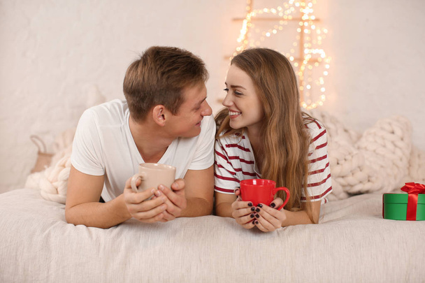 Liebendes junges Paar trinkt heiße Schokolade, während es auf dem Bett im weihnachtlich dekorierten Zimmer liegt - Foto, Bild