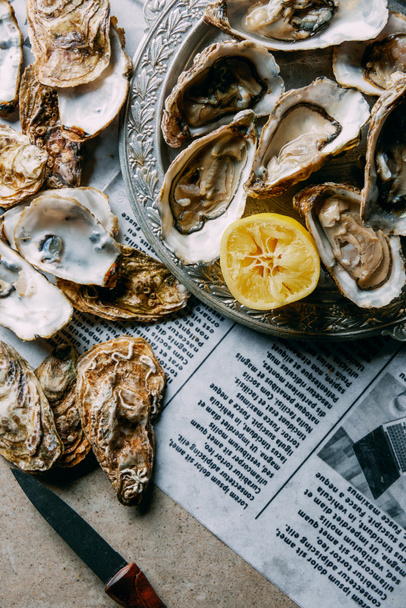 vlakke leggen met oesters, citroen en mes op krant op grijze oppervlak - Foto, afbeelding