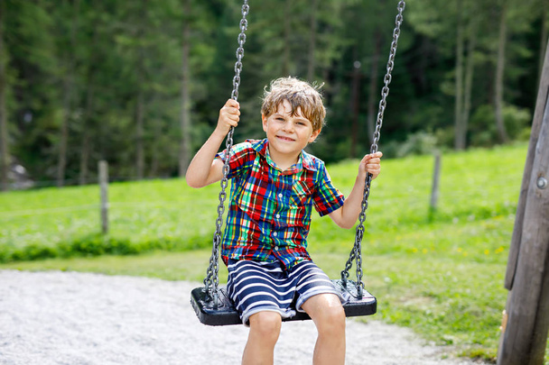 Zabawny chłopiec bawiący się huśtawką łańcuchową na placu zabaw na świeżym powietrzu, będąc mokrym rozbryzganym wodą. kołyszące się dziecko w letni dzień. Aktywny wypoczynek z dziećmi. Szczęśliwy płaczący chłopiec z kroplami deszczu na twarzy. - Zdjęcie, obraz