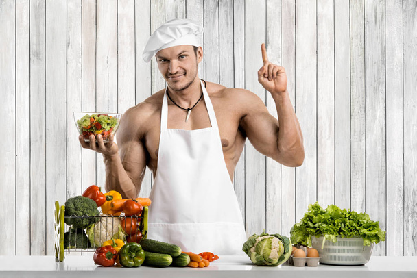Man bodybuilder on kitchen in white toque blanche and cook protective apron, concoction vegetables and fruit - Foto, imagen