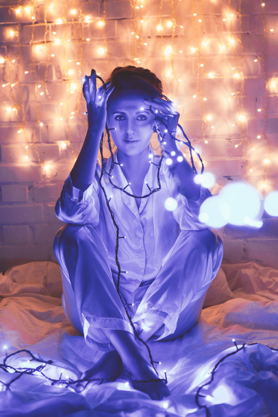 όμορφη γυναίκα στο πιτζάμες με γιορτινά φώτα κάθεται στο κρεβάτι στο σπίτι - Φωτογραφία, εικόνα