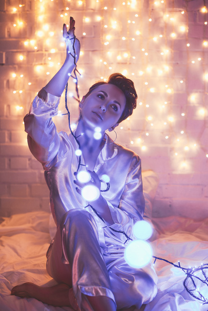woman in pajamas sitting on bed with christmas lights around - Photo, Image