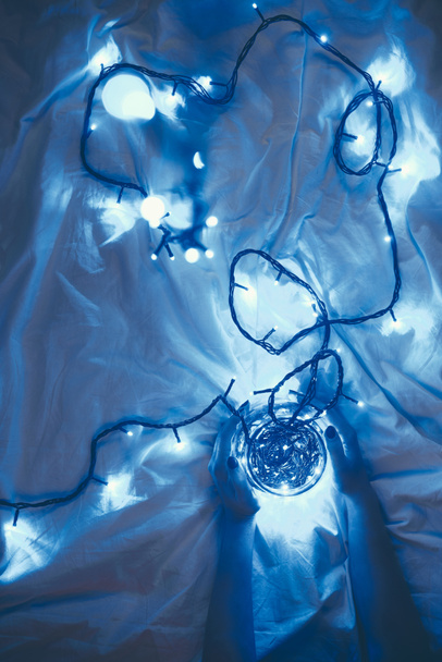 partial view of woman holding glass jar with blue christmas lights around on bed - Foto, imagen