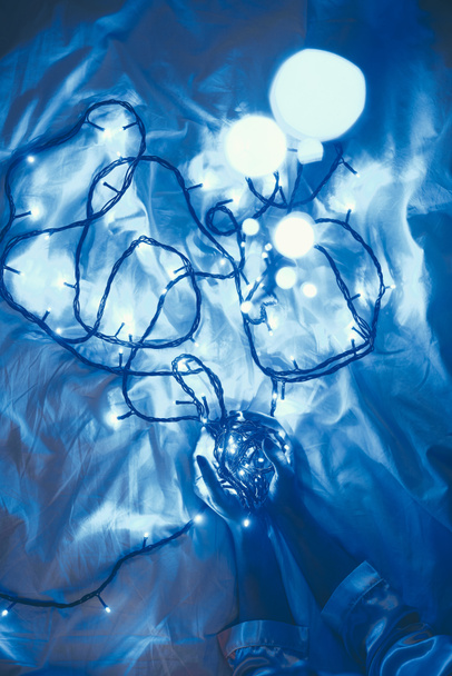 partial view of woman holding blue festive lights in hands on bed - Fotografie, Obrázek