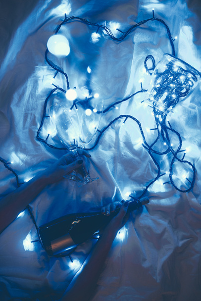 cropped shot of woman with bottle of champagne and glasses on bed with festive blue lights around - Photo, image