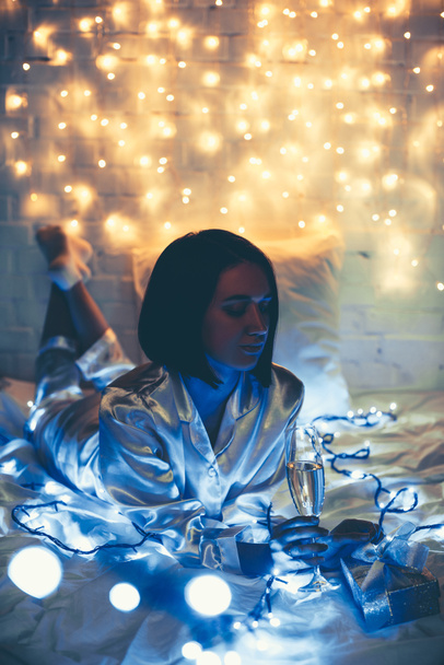 thoughtful woman in pajamas with glass of champagne and gift lying on bed with christmas lights around - Фото, зображення