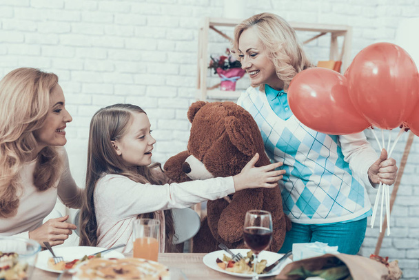 Grandmother Giving Toy Bear to Happy Girl at Home. Cake on Table. Happy Family. Mother with Daughter. Smiling Women. Smiling Grandmother. Celebration Concept. Toy Bear. Red Baloons. - Foto, Imagem