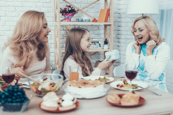 Family Celebrating Grandmother's Birthday at Home. Cake on Table. Happy Family. Mother with Daughter. Smiling Women. Smiling Grandmother. Celebration Concept. Glass of Wine. Giving Gift Box. - 写真・画像