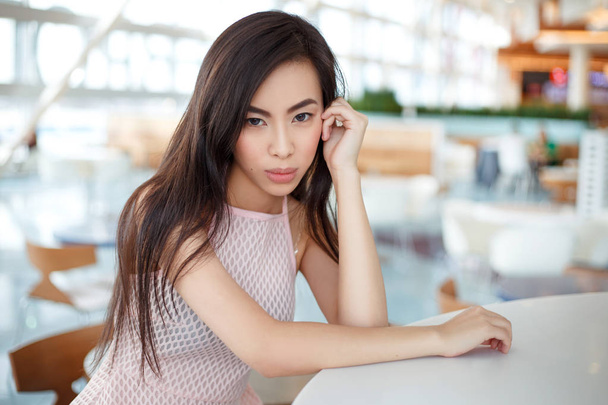 portrait of brunette young asian woman in pink dress sitting at table in cafe - Foto, afbeelding