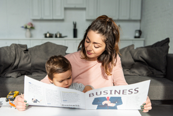 anya és kis fia olvasó teendő újság otthon a nappaliban - Fotó, kép