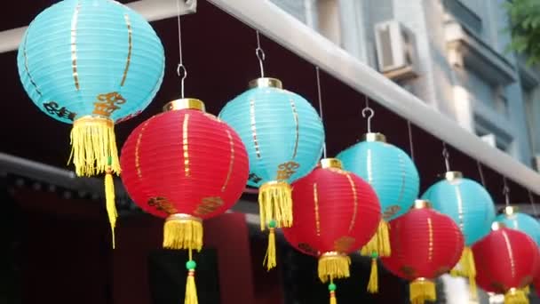 Chinese lantaarns zwaaien in de wind in de middag. Oosterse papier lichten van rode en blauwe kleuren zijn zwaaiend met de wind op het dak van een gebouw door dag op straat - Video