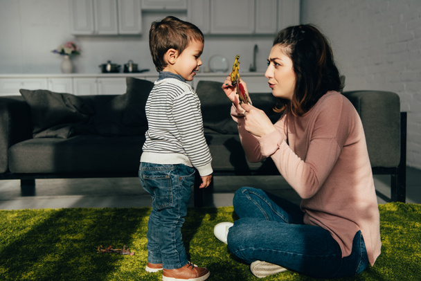 боковой вид матери, показывающей игрушечные обеды своему маленькому сыну в гостиной дома
 - Фото, изображение