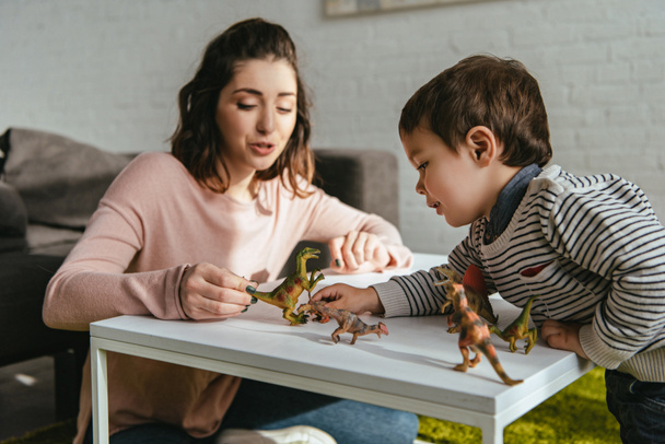 веселая женщина и маленький сын играют в игрушечных динозавров за столом в гостиной на дому
 - Фото, изображение