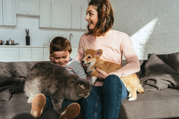 cheerful mother and son sitting on sofa with cat and dog in living room at home - Photo, Image