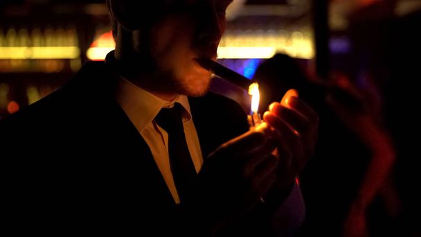 Businessman lighting cigar in elite bar, night atmosphere, dance floor, relax - Foto, imagen