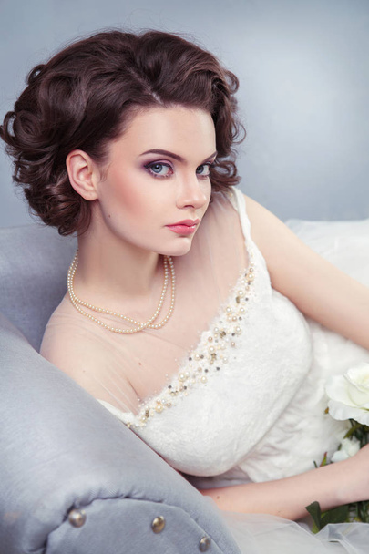 beautiful bride with elegant hairstyle in white wedding dress resting on sofa - Фото, изображение